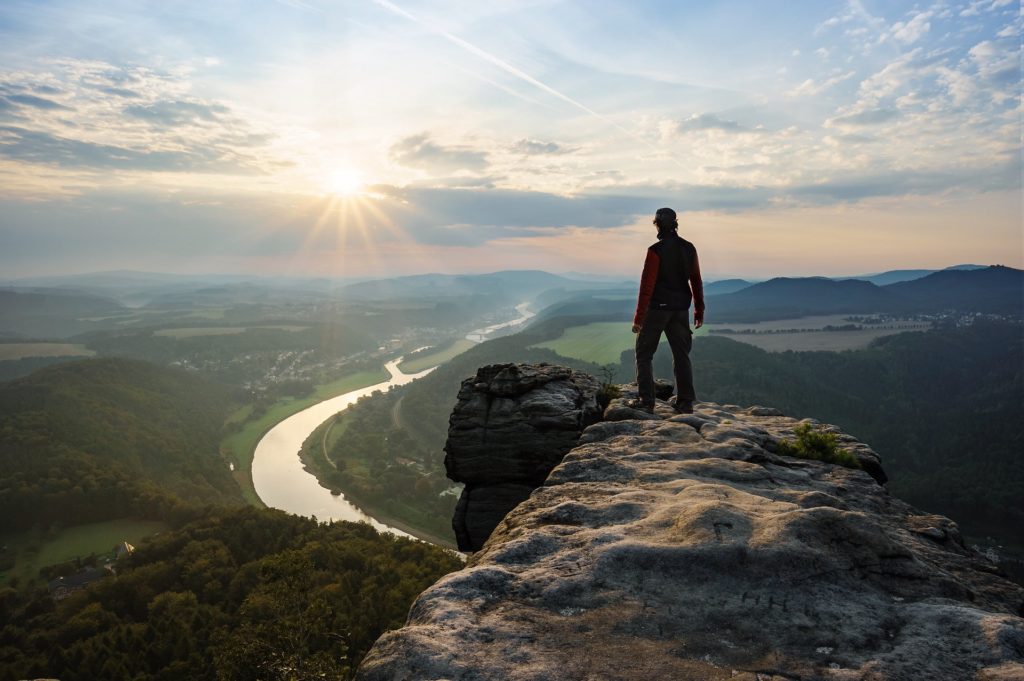 Bergsteiger auf Gipfel © hineswilliamjames Die Silhouette eines Bergsteigers im Sonnenuntergang, der am Rand eines Felsens steht und in das Tal mit einem Fluss hinab blickt.