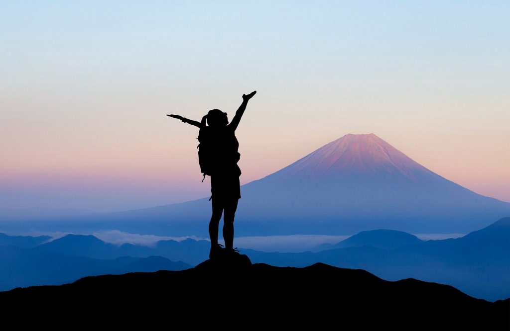 Reisender©Mohammed_hassan Eine reisende Frau steht auf einem Berg und streckt die Arme in den Himmel bei Sonnenaufgang mit einem Vulkan im Hintergrund
